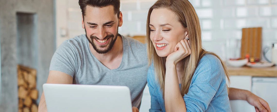 Eine Frau und ein Mann sitzen an einem Tisch und schauen auf einen Laptop. Beide lächeln. 