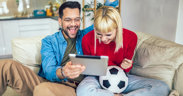 Ein Pärchen sitzt lachend auf einem Sofa und schaut sich auf dem Tablet ein Fußballspiel an